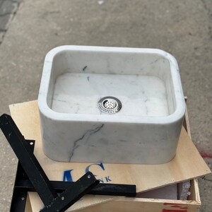 Close front view of a Carrara White marble vanity sink with a centered drain, deep hand-carved bowl, rounded corners and black steel mounting brackets.