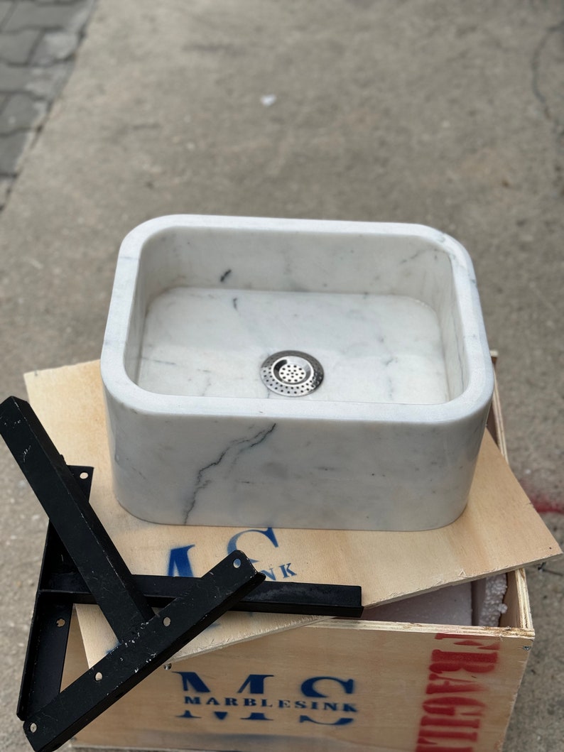 Close front view of a Carrara White marble vanity sink with a centered drain, deep hand-carved bowl, rounded corners and black steel mounting brackets.