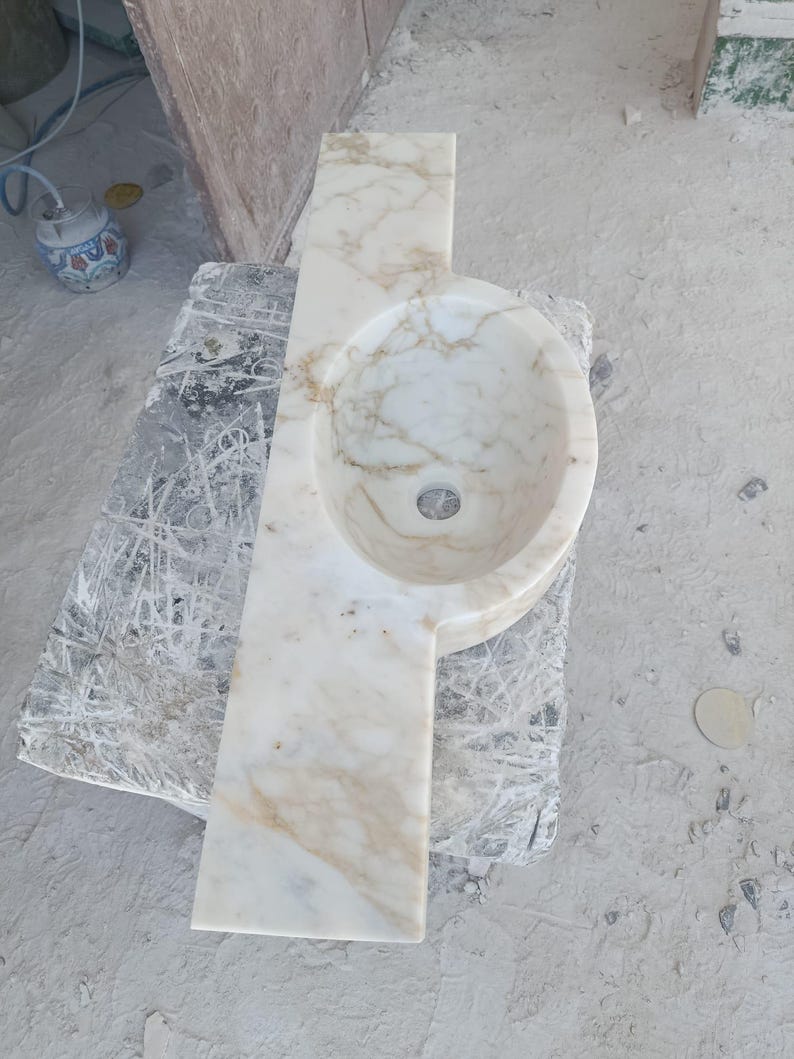 Luxurious wall-mounted Calacatta marble sink carved from a single block. Featuring 3 faucet holes, fluted front design, and floating vanity look. Ideal for modern, minimalist bathrooms. Custom sizes available. Handmade from natural Italian stone. Bathroom sink, marble vanity, stone basin, designer sink, fluted marble, wall mount sink, handmade sink, rectangular sink, luxury bathroom decor.
