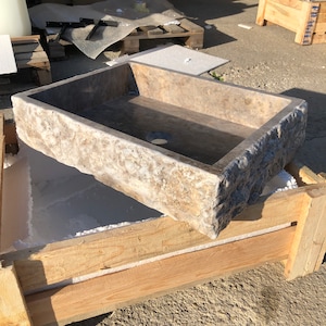 Op de afbeelding: Een rechthoekige stenen gootsteen met een ruw, getextureerd exterieur. De gootsteen is gemaakt van natuursteen en heeft een lichtbruine kleur met donkerbruine strepen. De gootsteen staat in een houten kist.