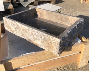 Puede incluir: Un lavabo rectangular de piedra con un exterior rugoso y texturizado. El lavabo está hecho de piedra natural y tiene un color marrón claro con vetas marrones más oscuras. El lavabo está sentado en una caja de madera.