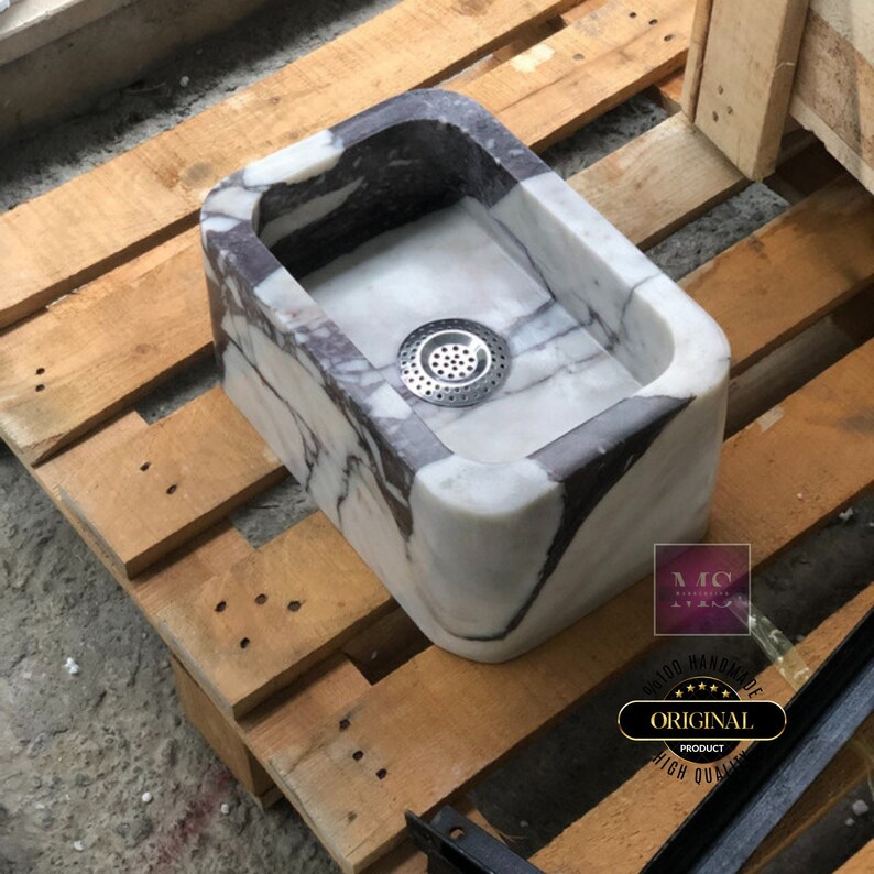 Hand-carved Calacatta Viola marble sink on a wooden pallet, showing the rectangular bowl, rounded corners and metal drain strainer.