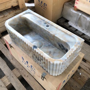 May include: A white marble sink with a fluted design. The sink has a rectangular shape and a single drain hole. The sink is sitting in a wooden crate.
