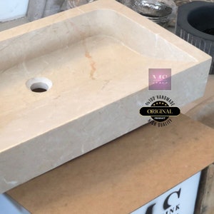 May include: A rectangular, off-white marble sink with a single drain hole. The sink is handmade and features the text "100% Handmade Original Product High Quality" in a circle.
