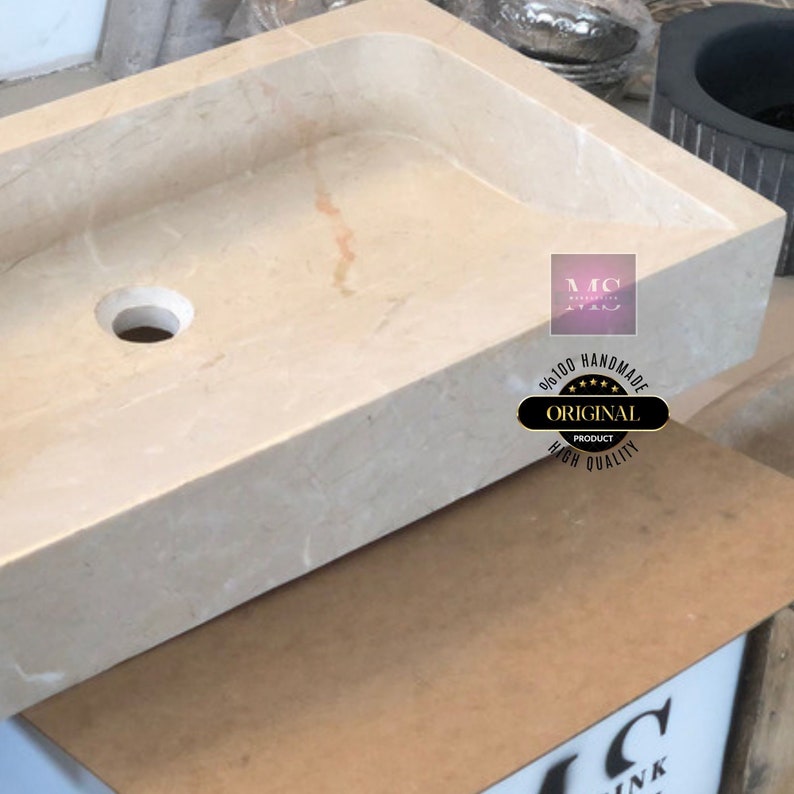 May include: A rectangular, off-white marble sink with a single drain hole. The sink is handmade and features the text "100% Handmade Original Product High Quality" in a circle.