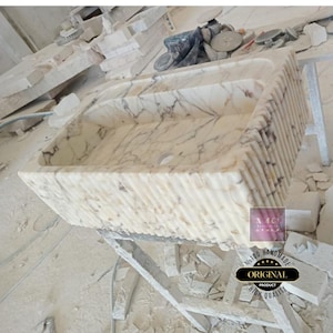 May include: A white marble sink with a rippled edge. The sink is handmade and has a high quality finish. The sink is sitting on a wooden stand.