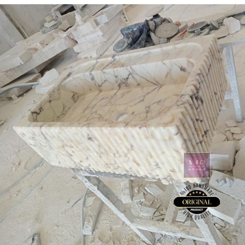 May include: A white marble sink with a rippled edge. The sink is handmade and has a high quality finish. The sink is sitting on a wooden stand.