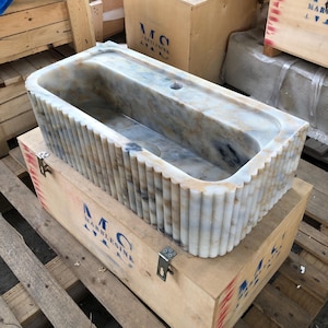 May include: A white marble sink with a fluted design. The sink is in a wooden crate with the text "MC MARBLESINK" printed on the side.