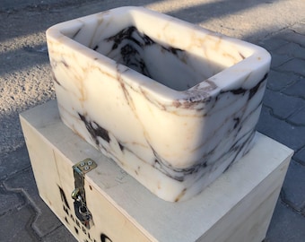 May include: A rectangular white marble planter with brown and gold veining. The planter is sitting on a wooden crate.