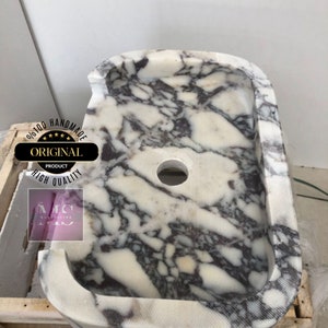 May include: A white marble sink with a gray and purple marbling pattern. The sink has a rounded rectangular shape with a single drain hole in the center. The sink is handmade and is 100% original.
