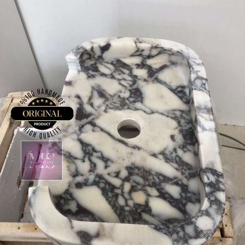 May include: A white marble sink with a gray and purple marbling pattern. The sink has a rounded rectangular shape with a single drain hole in the center. The sink is handmade and is 100% original.