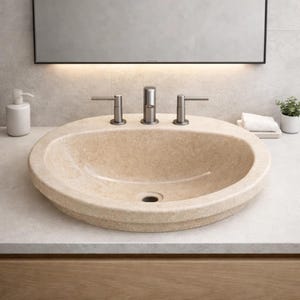 May include: A beige oval bathroom sink with a brushed metal faucet and handles. The sink is set into a light-colored countertop. A white soap dispenser and a small potted plant are visible in the background.