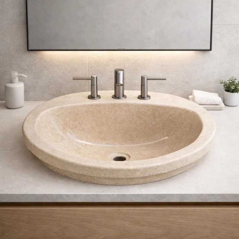 May include: A beige oval bathroom sink with a brushed metal faucet and handles. The sink is set into a light-colored countertop. A white soap dispenser and a small potted plant are visible in the background.
