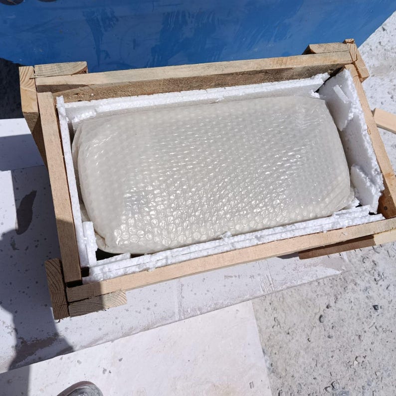 May include: A rectangular package wrapped in bubble wrap and foam padding, inside a wooden crate. The crate is made of light-colored wood and is open at the top, revealing the contents. The background is a mix of concrete and a blue surface.