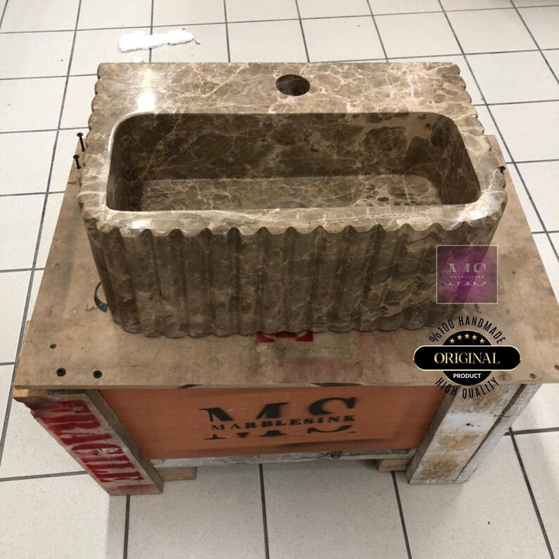 May include: A brown marble sink with a rippled edge, sitting on a wooden crate. The crate is labeled "MC MARBLESINK". The sink is handmade and is a unique piece of bathroom decor.