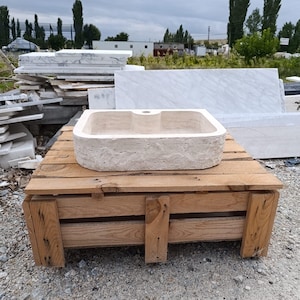 Op de afbeelding: Een rechthoekige beige stenen wastafel met een ruwe, getextureerde buitenkant en een gladde, gepolijste binnenkant. De wastafel heeft een enkel gecentreerd kraangat aan de achterrand en een natuurlijke, poreuze uitstraling van travertijn.