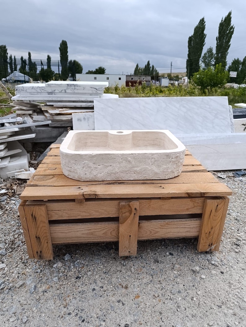 Op de afbeelding: Een rechthoekige beige stenen wastafel met een ruwe, getextureerde buitenkant en een gladde, gepolijste binnenkant. De wastafel heeft een enkel gecentreerd kraangat aan de achterrand en een natuurlijke, poreuze uitstraling van travertijn.