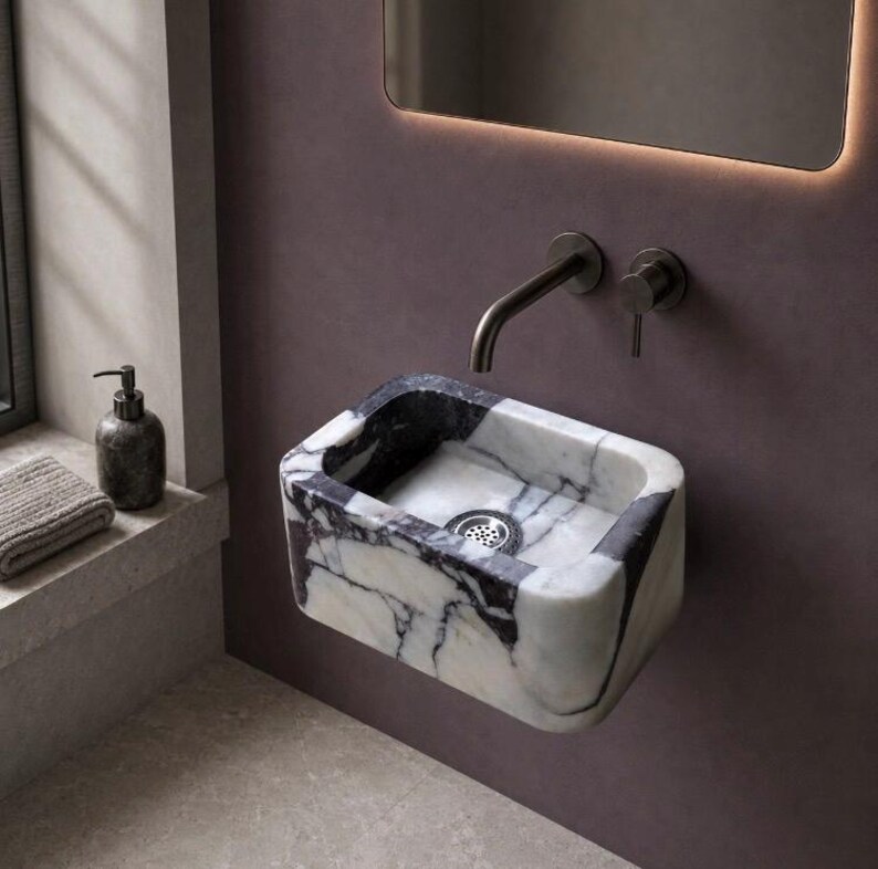 Wall-mounted Calacatta Viola marble sink in a modern burgundy powder room with a brass faucet, illuminated mirror and warm lighting.