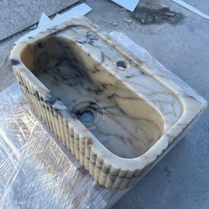 "Handmade Calacatta marble wall-mounted sink with fluted front design, shown in a minimalist bathroom setting. Featuring a single faucet hole and rear shelf for soap or accessories. Perfect for modern, luxury interiors. Custom sizes available. Polished interior, natural veining, and matte black faucet compatibility."