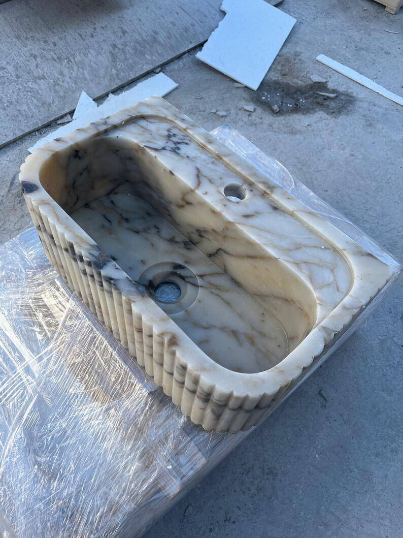 "Handmade Calacatta marble wall-mounted sink with fluted front design, shown in a minimalist bathroom setting. Featuring a single faucet hole and rear shelf for soap or accessories. Perfect for modern, luxury interiors. Custom sizes available. Polished interior, natural veining, and matte black faucet compatibility."