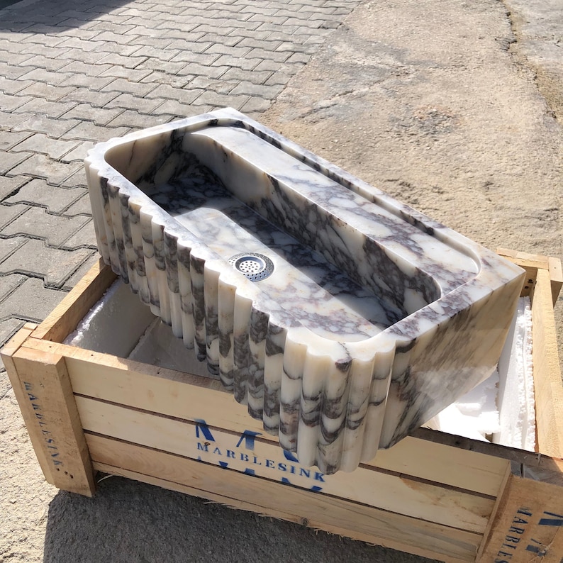Hand-carved fluted Calacatta Viola marble sink displayed on a protective wooden shipping crate in the stone workshop.