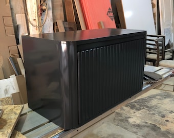 May include: A dark grey cabinet with a black slatted door. The cabinet has a rounded edge on the front and is sitting on a wooden surface.