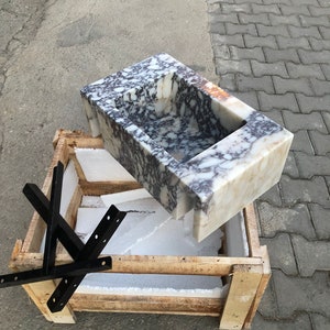 May include: A white marble sink with purple and gray veining is packaged in a wooden crate with foam padding. The sink is rectangular with a rounded edge and a small rectangular cutout in the center.