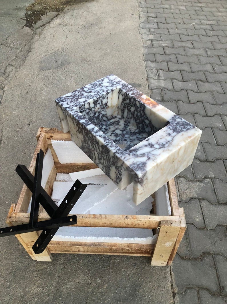 May include: A white marble sink with purple and gray veining is packaged in a wooden crate with foam padding. The sink is rectangular with a rounded edge and a small rectangular cutout in the center.