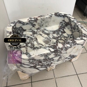 May include: A white marble sink with purple and gray veining. The sink is oval shaped and has a single drain hole. The sink is handmade and is 100% original.