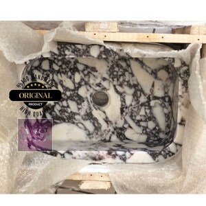 May include: A white marble sink with purple and gray veining. The sink has a round drain hole and a slightly curved edge. The sink is handmade and is 100% original.