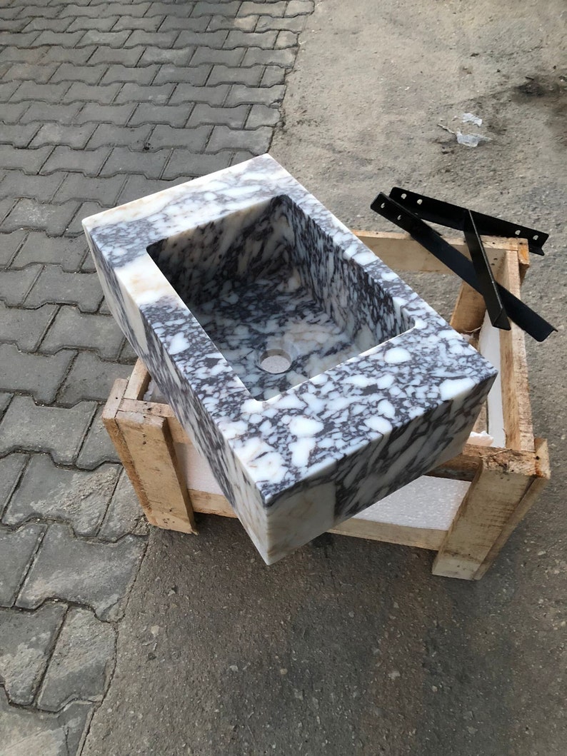 May include: A white marble sink with gray veining, sitting on a wooden crate. The sink is rectangular and has a rounded edge.