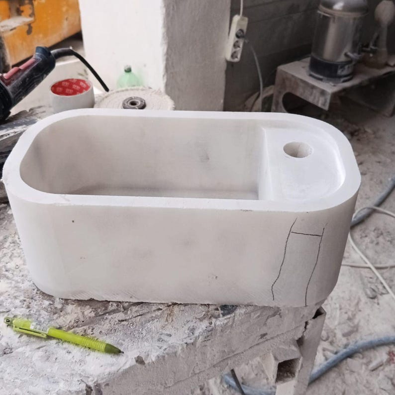 May include: A rectangular, off-white concrete sink with rounded edges and a built-in soap dish. The sink is unfinished, showing the raw texture of the material. A green pen rests on the surface in the foreground.