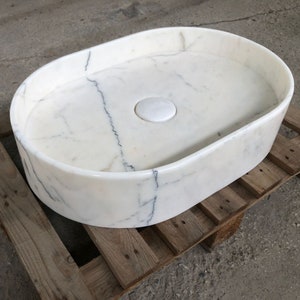 Może przedstawiać: Biała owalna umywalka łazienkowa z marmuru z otworem odpływowym i białym korkiem ceramicznym. Umywalka stoi na drewnianej palecie.