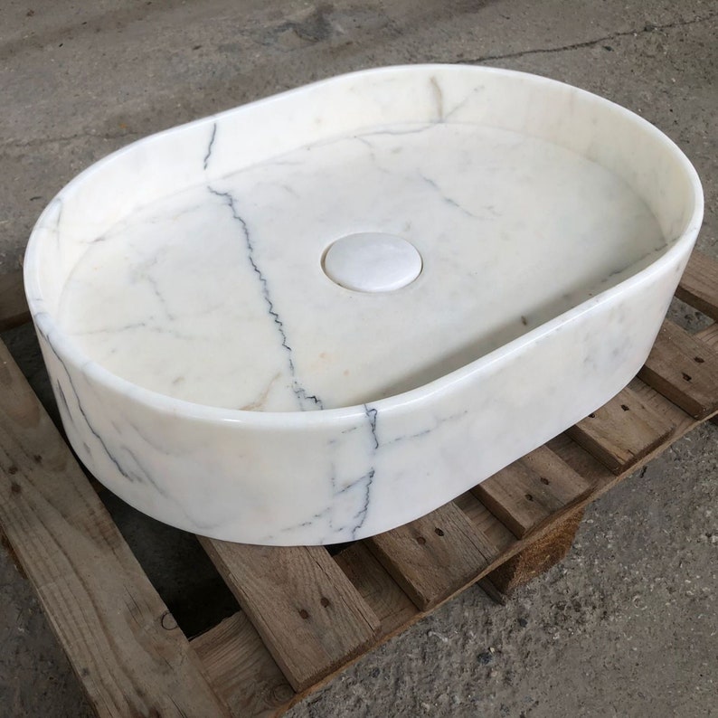 Może przedstawiać: Biała owalna umywalka łazienkowa z marmuru z otworem odpływowym i białym korkiem ceramicznym. Umywalka stoi na drewnianej palecie.