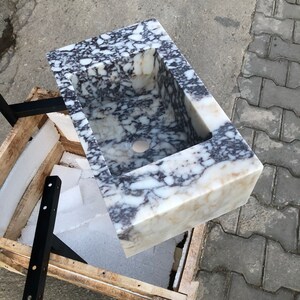 May include: A white marble sink with black and gray veining. The sink is rectangular and has a square cutout for the drain. The sink is sitting on a wooden crate.
