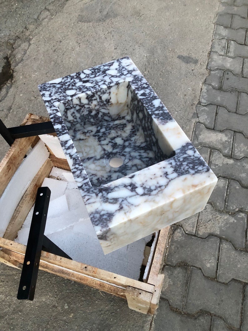 May include: A white marble sink with black and gray veining. The sink is rectangular and has a square cutout for the drain. The sink is sitting on a wooden crate.