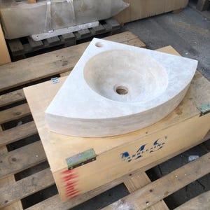 May include: A white corner sink made of travertine stone. The sink is sitting on a wooden pallet with a blue and white logo that says "M.C. MARBLE & STONE".