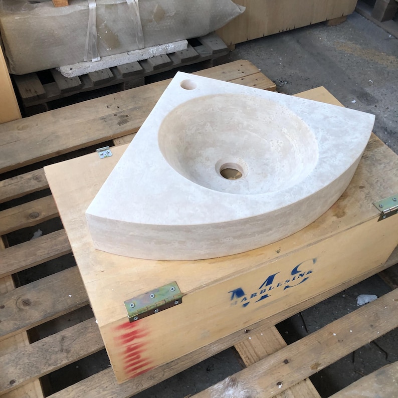 May include: A white corner sink made of travertine stone. The sink is sitting on a wooden pallet with a blue and white logo that says "M.C. MARBLE & STONE".