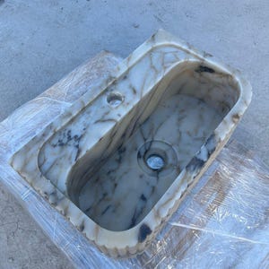 "Handmade Calacatta marble wall-mounted sink with fluted front design, shown in a minimalist bathroom setting. Featuring a single faucet hole and rear shelf for soap or accessories. Perfect for modern, luxury interiors. Custom sizes available. Polished interior, natural veining, and matte black faucet compatibility."