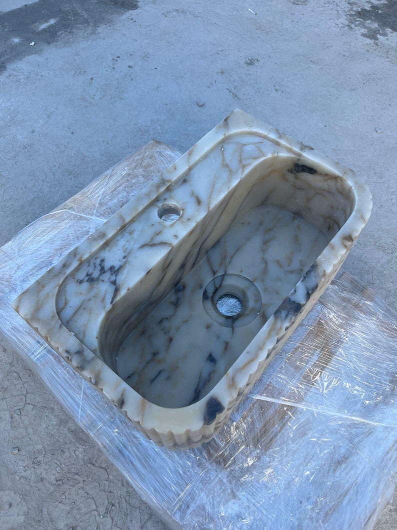 "Handmade Calacatta marble wall-mounted sink with fluted front design, shown in a minimalist bathroom setting. Featuring a single faucet hole and rear shelf for soap or accessories. Perfect for modern, luxury interiors. Custom sizes available. Polished interior, natural veining, and matte black faucet compatibility."