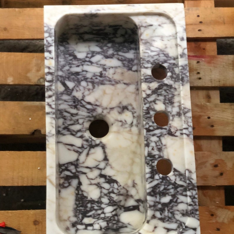 Puede incluir: Un lavabo de m&aacute;rmol blanco con un patr&oacute;n de vetas p&uacute;rpuras y grises. El lavabo tiene una forma rectangular con un borde redondeado y tres agujeros en el lado derecho.