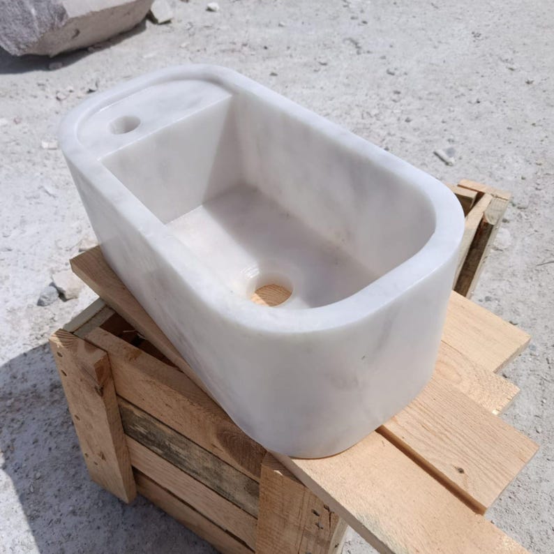 May include: A rectangular white marble sink with a rounded interior and a small rectangular shelf on the left side. The sink has a drain hole and a faucet hole. The sink is sitting on a wooden crate.