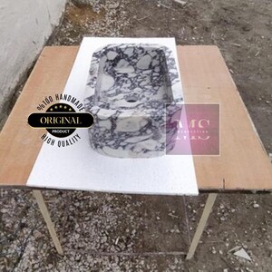 Op de afbeelding: Een witte marmeren gootsteen met paarse aderen, die op een houten tafel staat. De gootsteen is ovaalvormig en heeft een afvoergat in het midden. De tafel is van hout en heeft een wit oppervlak. De gootsteen is handgemaakt en 100% origineel.