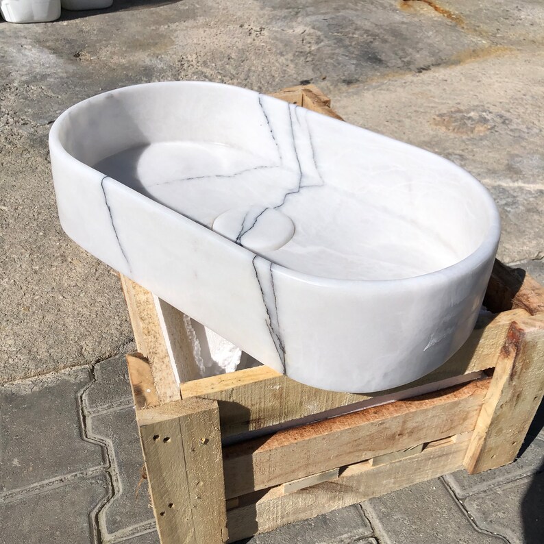 Puede incluir: Lavabo ovalado de m&aacute;rmol blanco con vetas grises, sentado en una caja de madera.