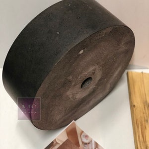 Pu&ograve; includere: Un vaso da fiori in cemento grigio scuro con una texture ruvida e un piccolo foro al centro. Il vaso &egrave; rotondo e ha una base piatta.