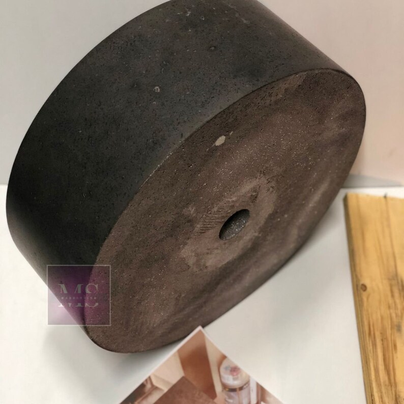 Pu&ograve; includere: Un vaso da fiori in cemento grigio scuro con una texture ruvida e un piccolo foro al centro. Il vaso &egrave; rotondo e ha una base piatta.