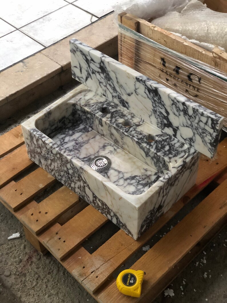 May include: A white marble sink with a grey veining pattern. The sink is rectangular and has a single drain. The sink is sitting on a wooden pallet.