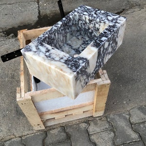 May include: A white marble sink with black and brown veining. The sink is rectangular and has a square opening. The sink is sitting on a wooden crate.