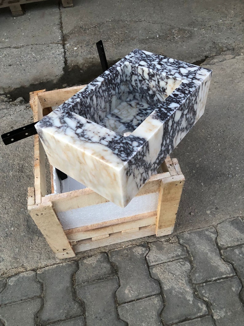May include: A white marble sink with black and brown veining. The sink is rectangular and has a square opening. The sink is sitting on a wooden crate.