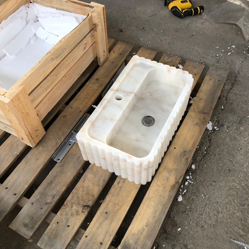 May include: A white marble sink with a rippled edge sits on a wooden pallet. The sink has a single drain hole and a small overflow drain.